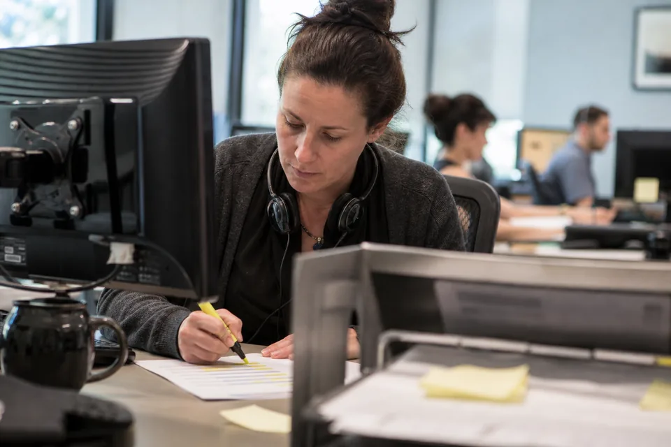 Customer experience lead at a support floor desk marking a printed one-page survey summary with a highlighter, headset around her neck
